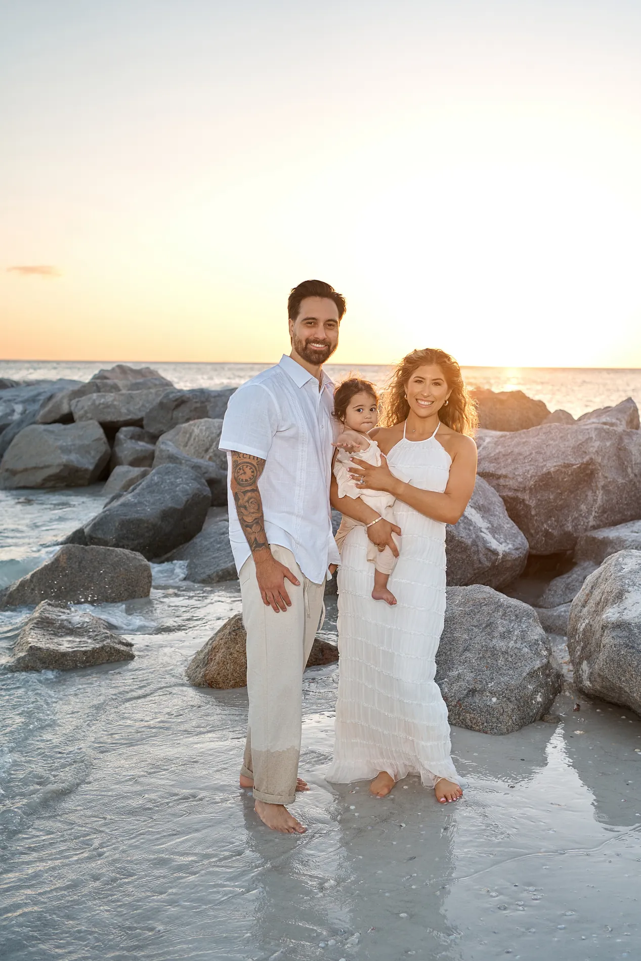 A young couple standing in shallow water beside big rocks, the father holding their baby, a warm sunset sky behind them