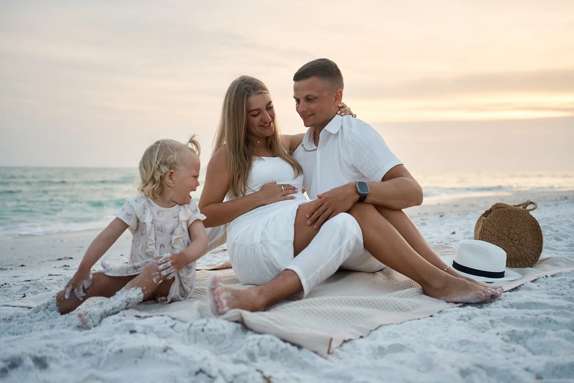 Parents in white sitting on a blanket on the white sand of South Lido after sunset, the mother resting a hand on her belly, their little daughter laughing beside them with sand on her legs