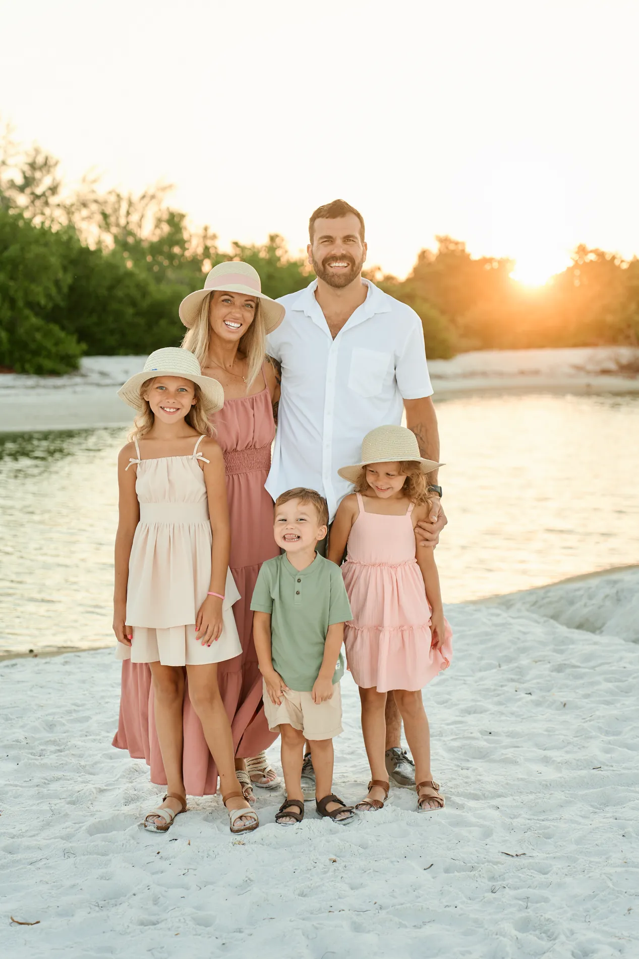 The same family of five standing close together by the water early in the morning: the mother in a dusty rose maxi dress and straw hat, the father in a white shirt, the girls in pink and cream dresses and straw hats, the boy in a sage shirt