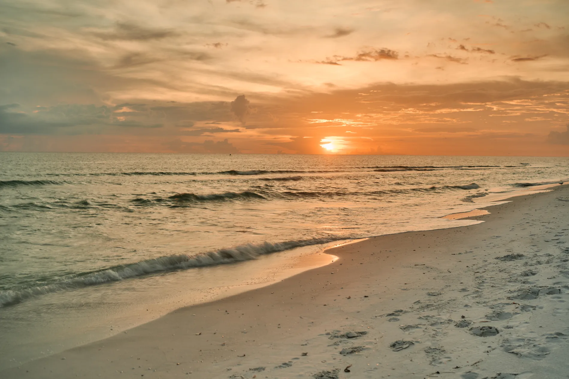 An empty beach on South Lido at sunset, the sun dropping through a narrow gap between a bank of cloud and the Gulf, the sky in orange and grey bands over soft waves