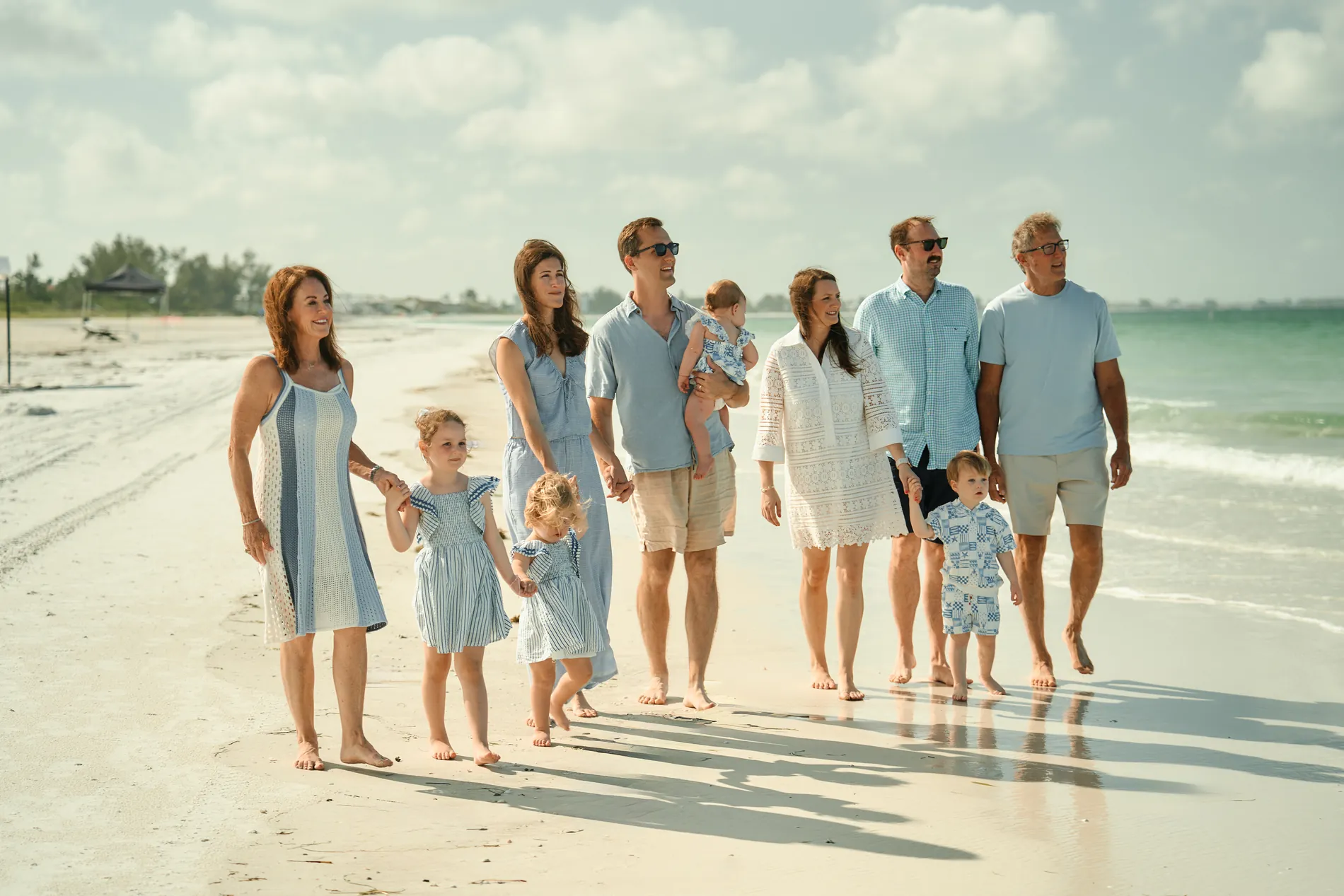 A big family in pale blue and white, babies in arms and small children by the hand, walking toward the camera along the white sand