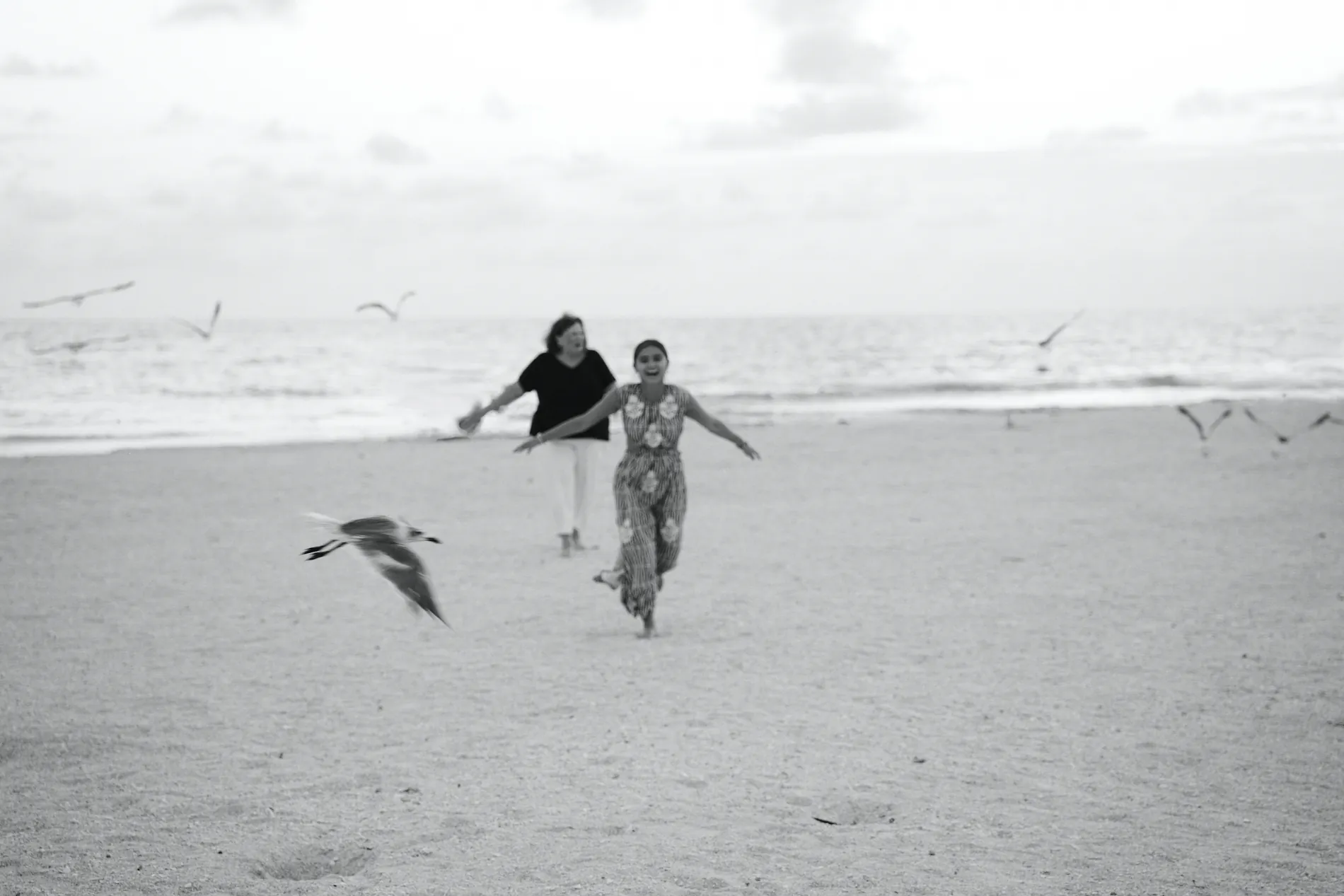 A black and white photograph of a mother and her grown daughter running across the sand on Lido Key, gulls flying around them
