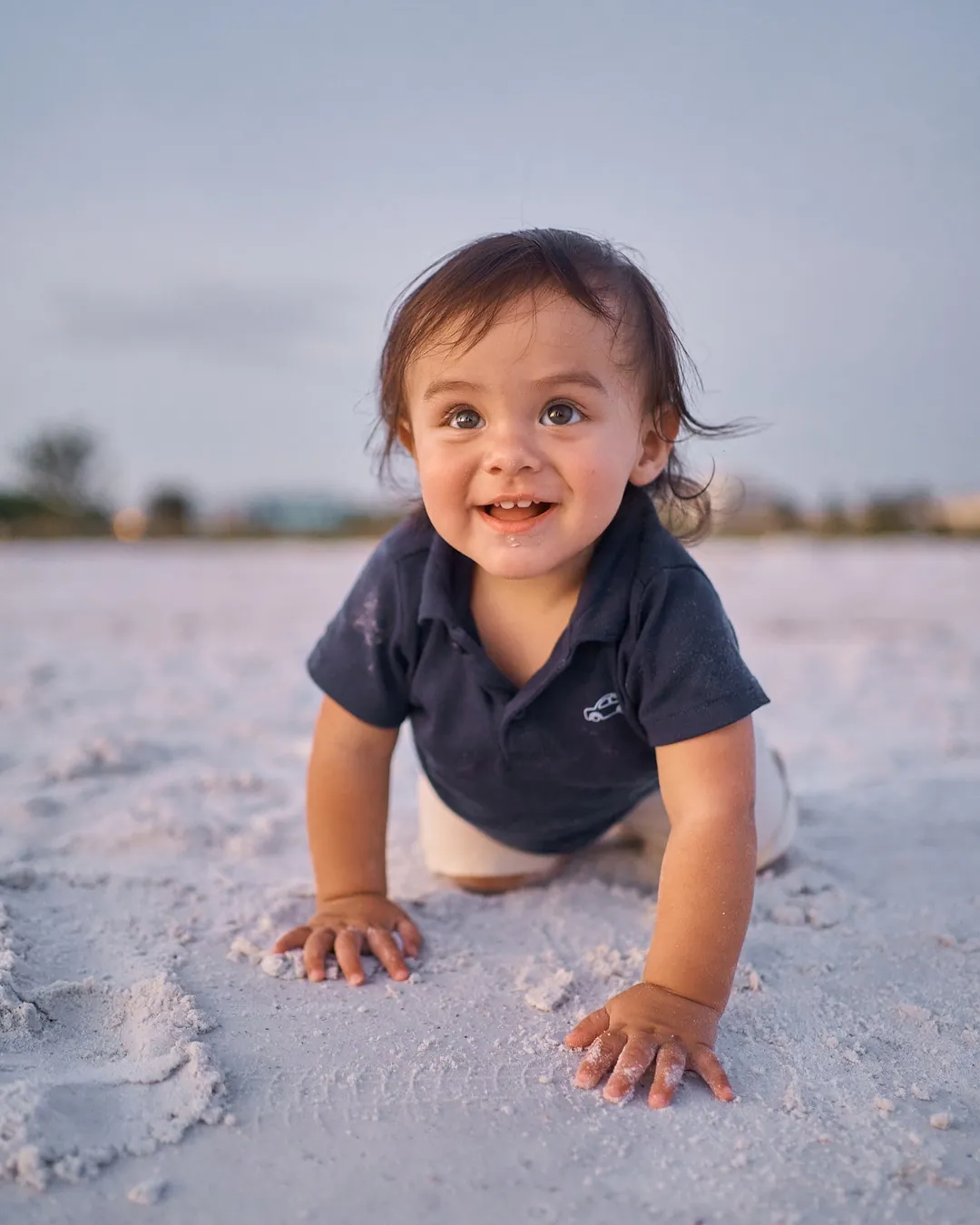 A baby boy in a navy shirt crawling across the white sand of Siesta Key at dusk, smiling at the camera