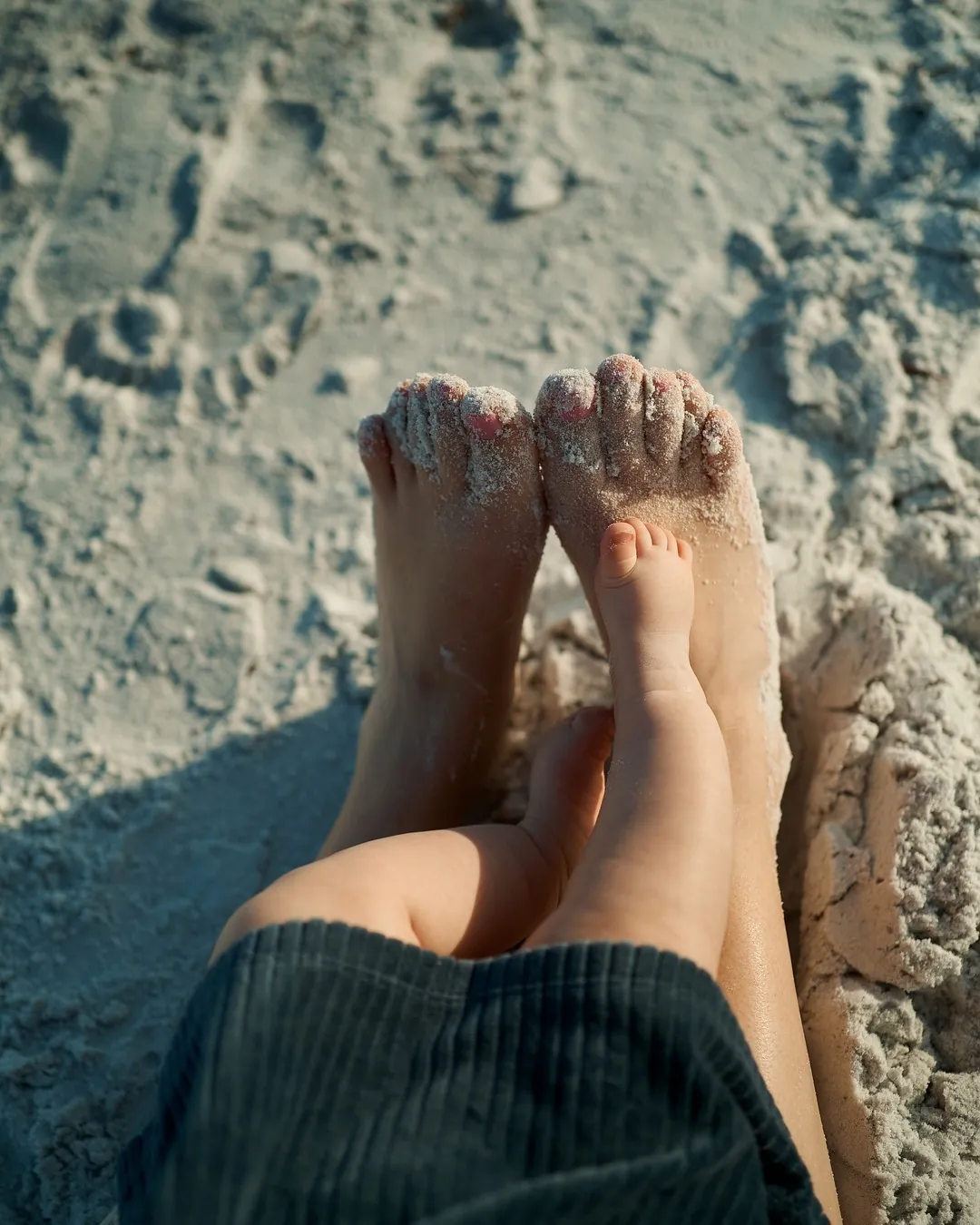 A baby's sandy feet resting on a parent's legs on the white sand of Siesta Key