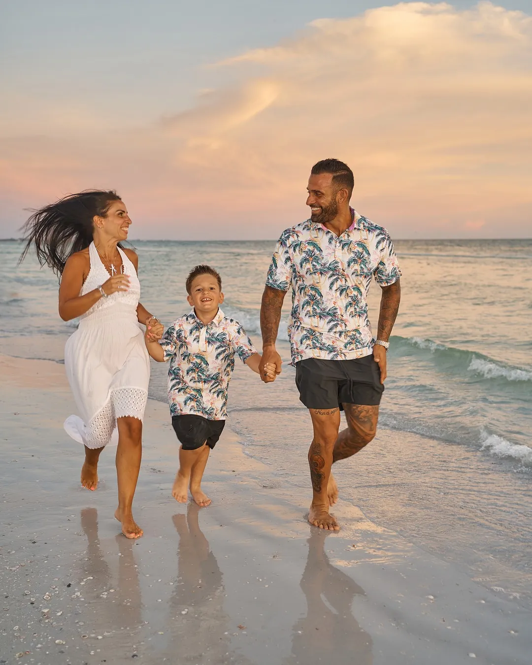 Parents running along the wet sand of Siesta Key with their small son between them, a pink sunset sky behind