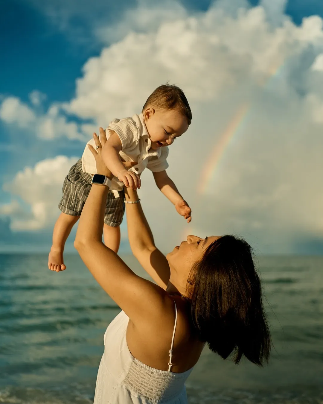 A mother lifting her laughing baby son above her head on Siesta Key, a rainbow in the clouds over the Gulf behind them