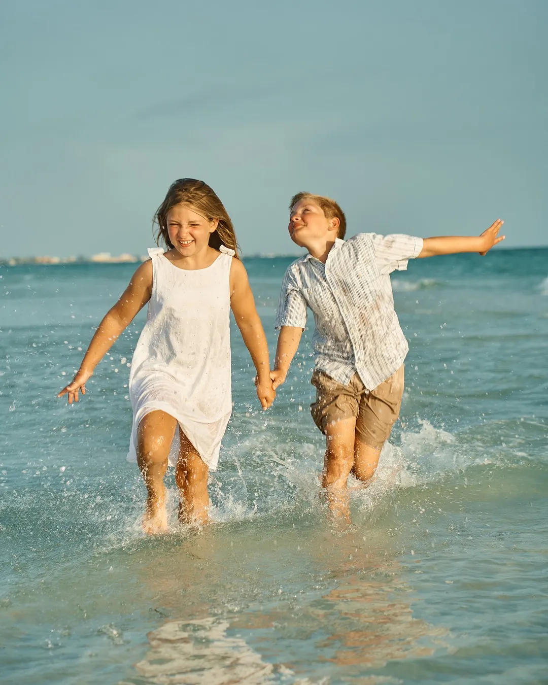 A sister and brother holding hands and running through the shallow turquoise water on Siesta Key, spray flying around them