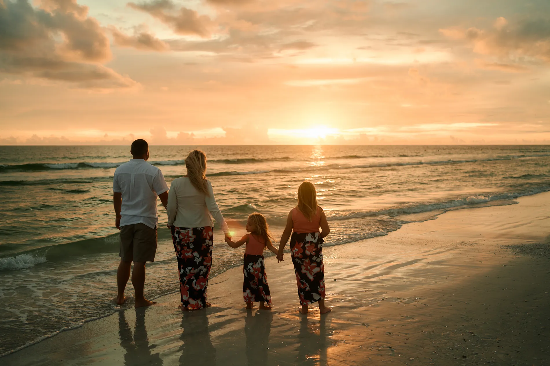 A family of four seen from behind, holding hands at the edge of the water on Siesta Key and watching the sun set over the Gulf