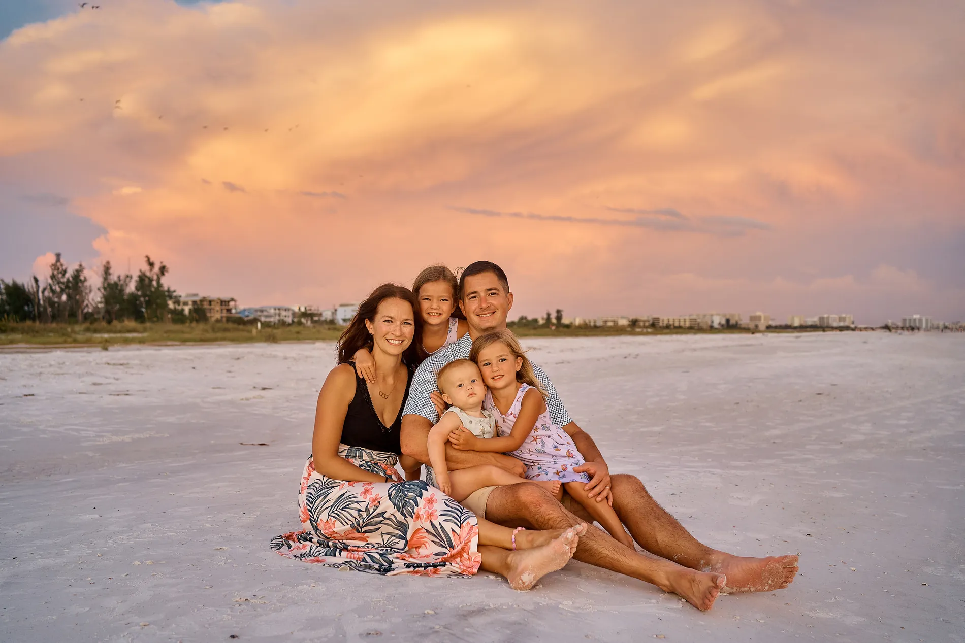 A family of five sitting close together on the white sand of Siesta Key under a pink evening sky