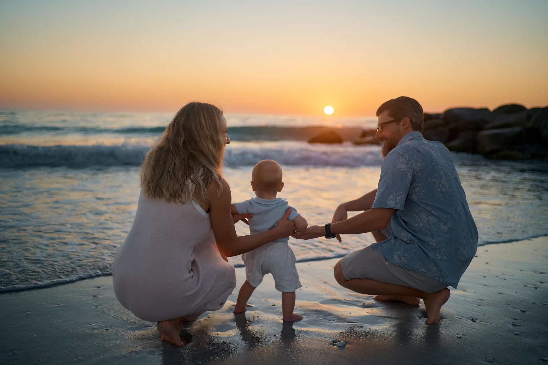 A toddler taking steps between his crouching parents at the edge of the water, the sun setting over the Gulf behind them
