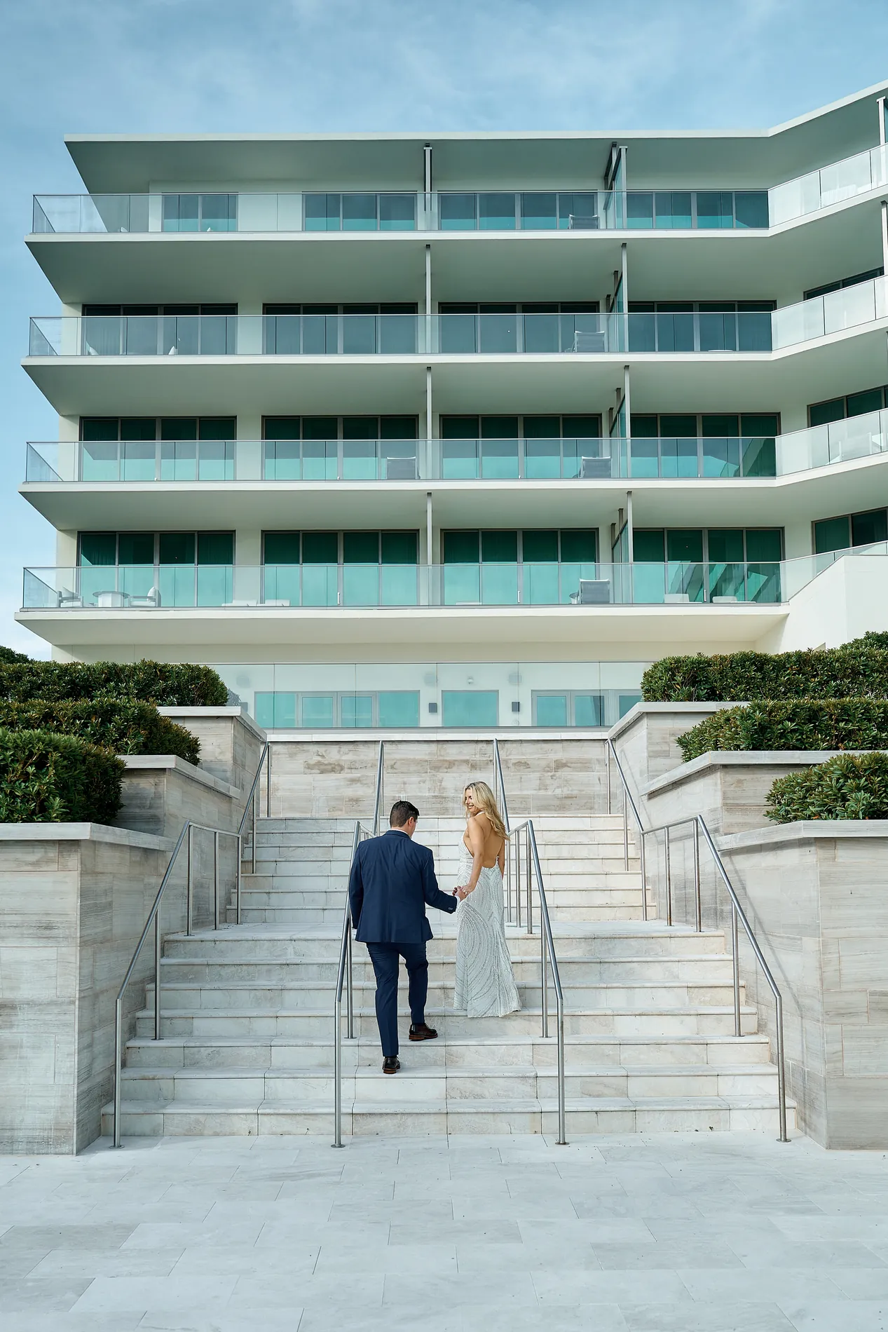 A couple in a navy suit and a beaded gown walking hand in hand up the wide stone stairs toward the white tower of The St. Regis Longboat Key