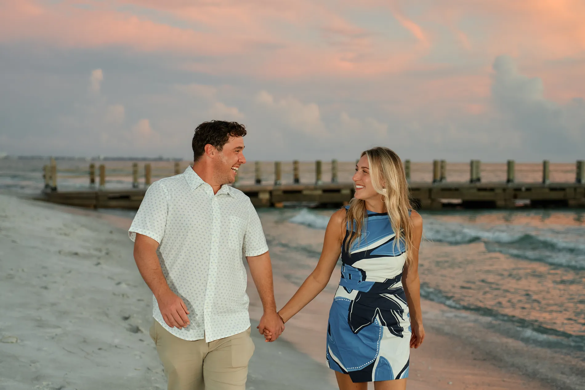 A newly engaged couple walking hand in hand along the water at The St. Regis Longboat Key, the pier behind them under a pink sky after sunset