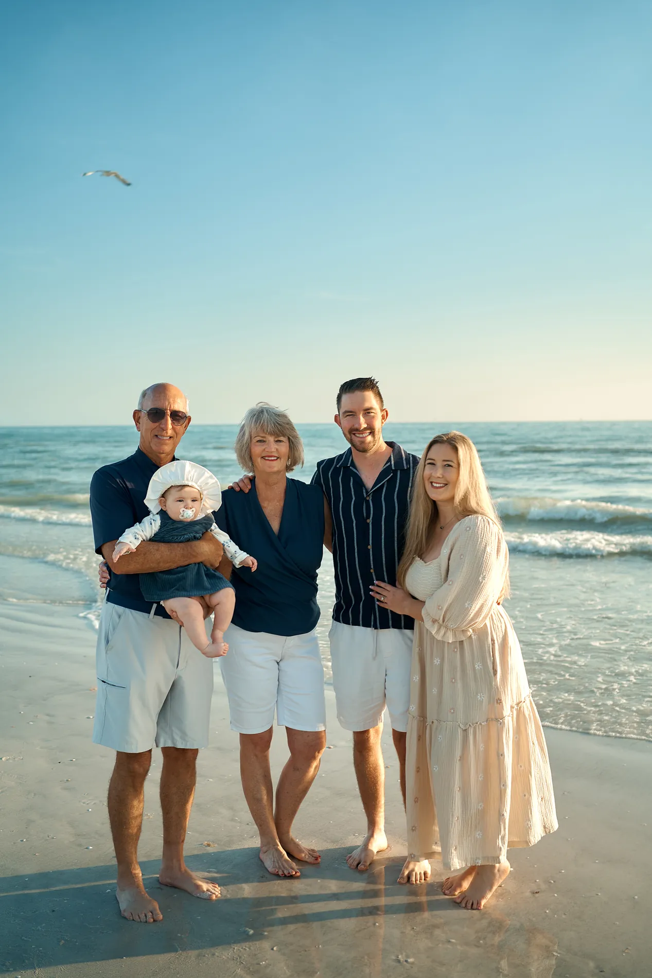 Grandparents holding their baby granddaughter in a white bonnet beside their son and his wife at the edge of the water, everyone in navy and white, a gull in the clear sky above them