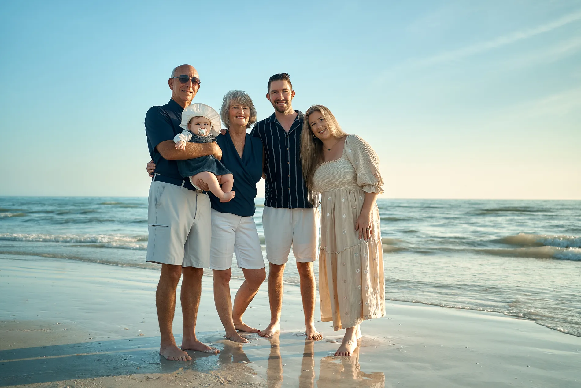 Grandparents holding their grandson beside his young parents at the edge of the water, under a clear blue sky