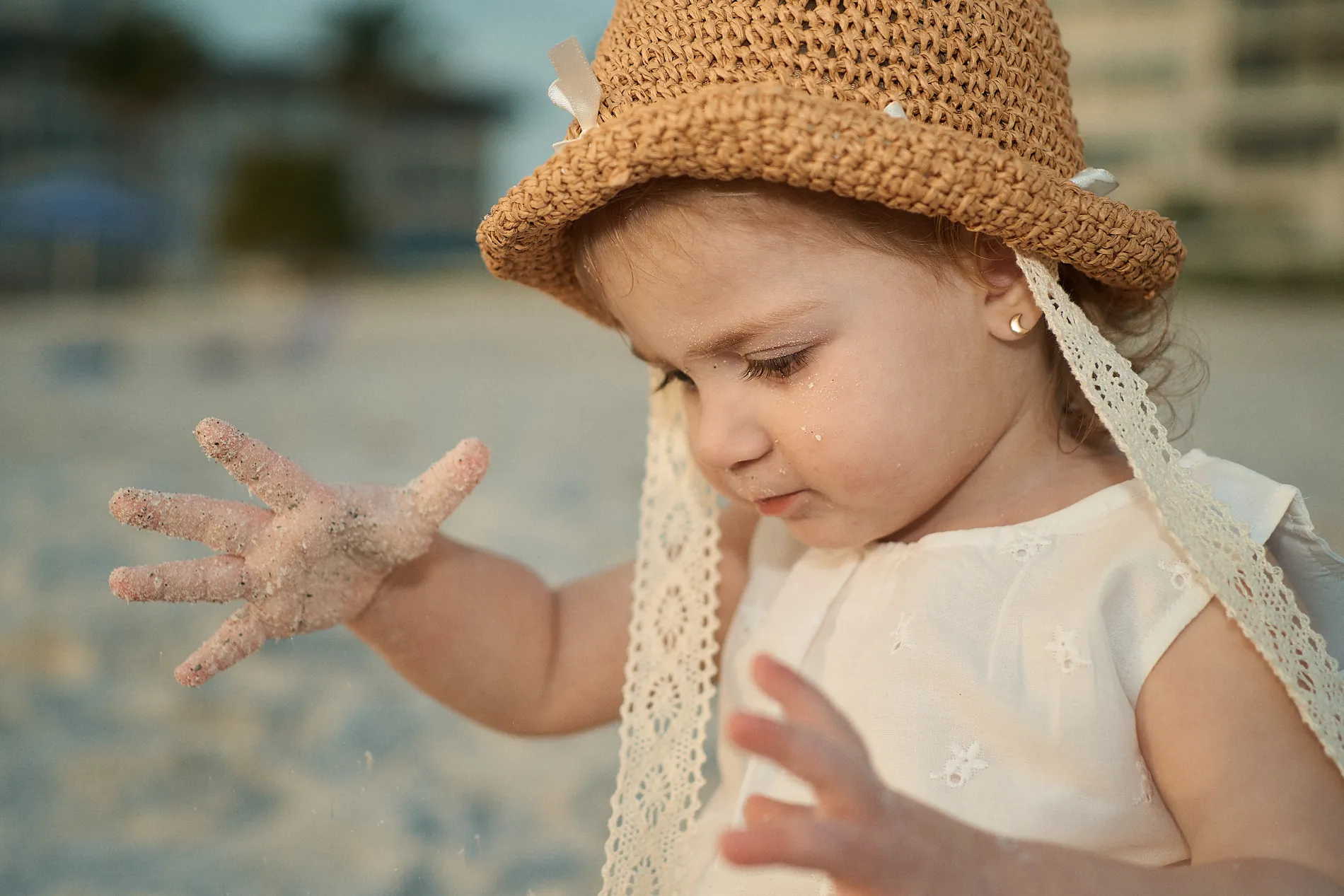 A little girl in a straw hat with lace ties and a white embroidered blouse studying her sandy hand