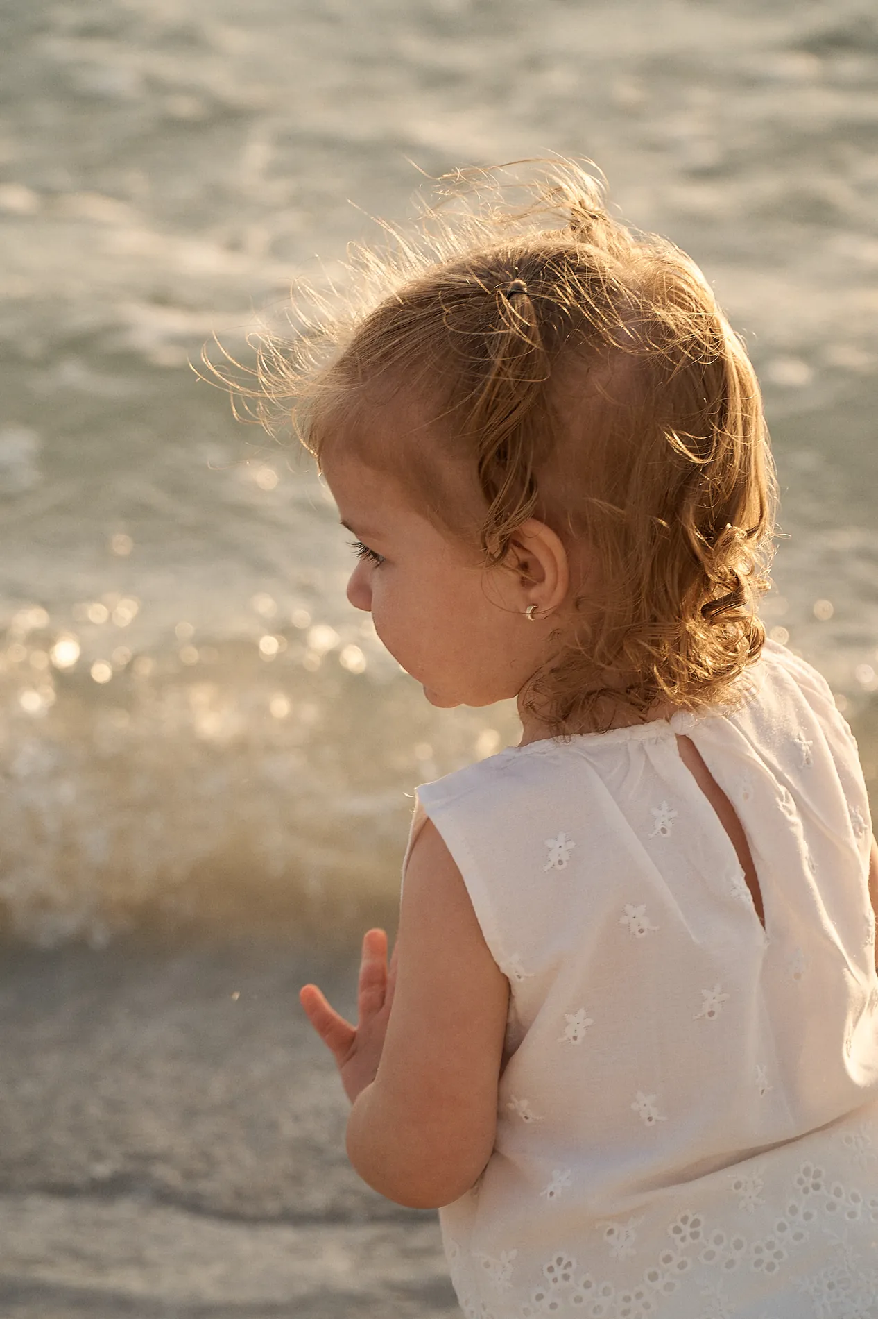 A curly-haired toddler girl in a white eyelet dress at the edge of the water, the low evening sun sparkling on the small waves behind her