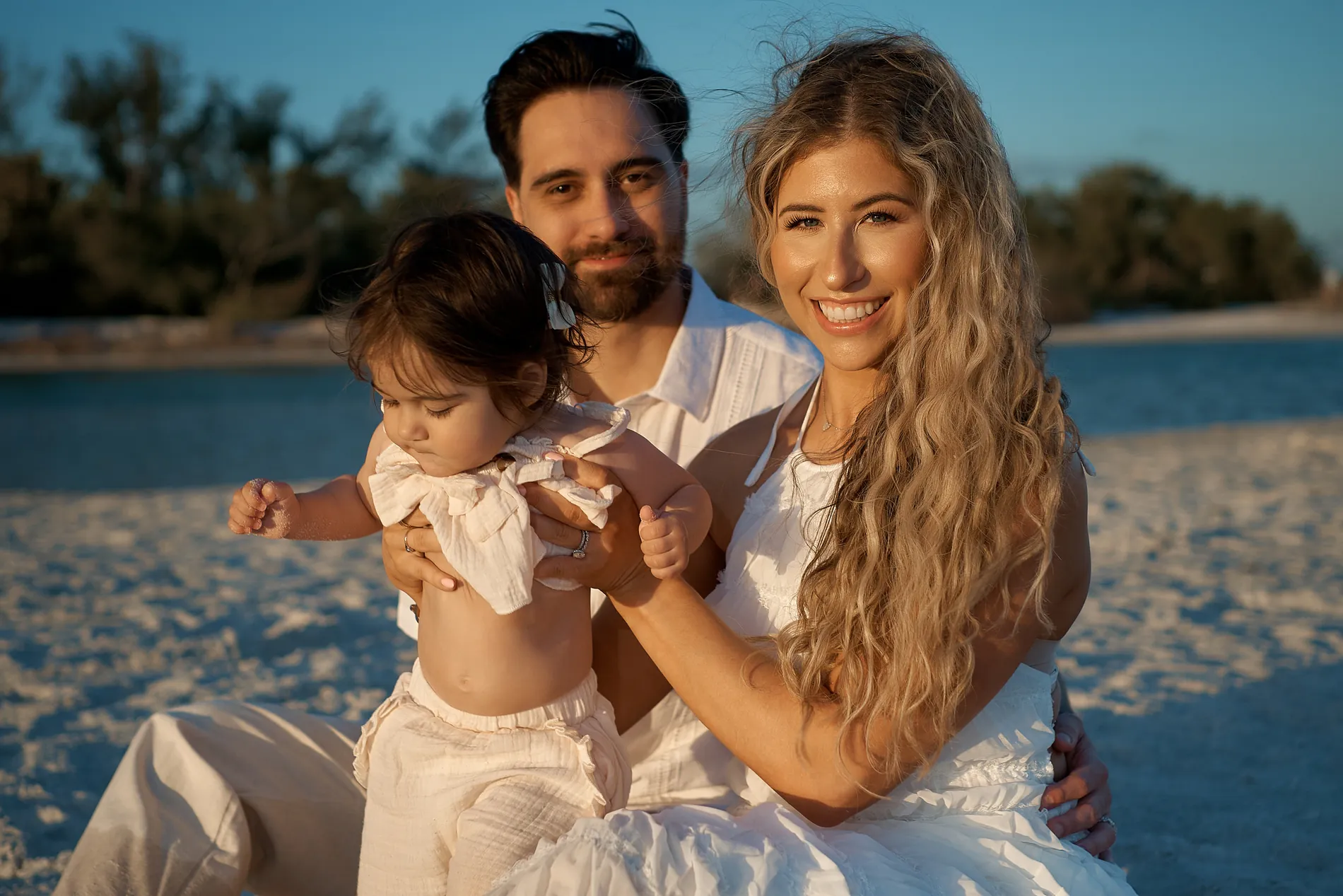 A mother, father and baby girl all in white sitting on the sand, photographed low so that dark blue water and green trees are behind them instead of the sand