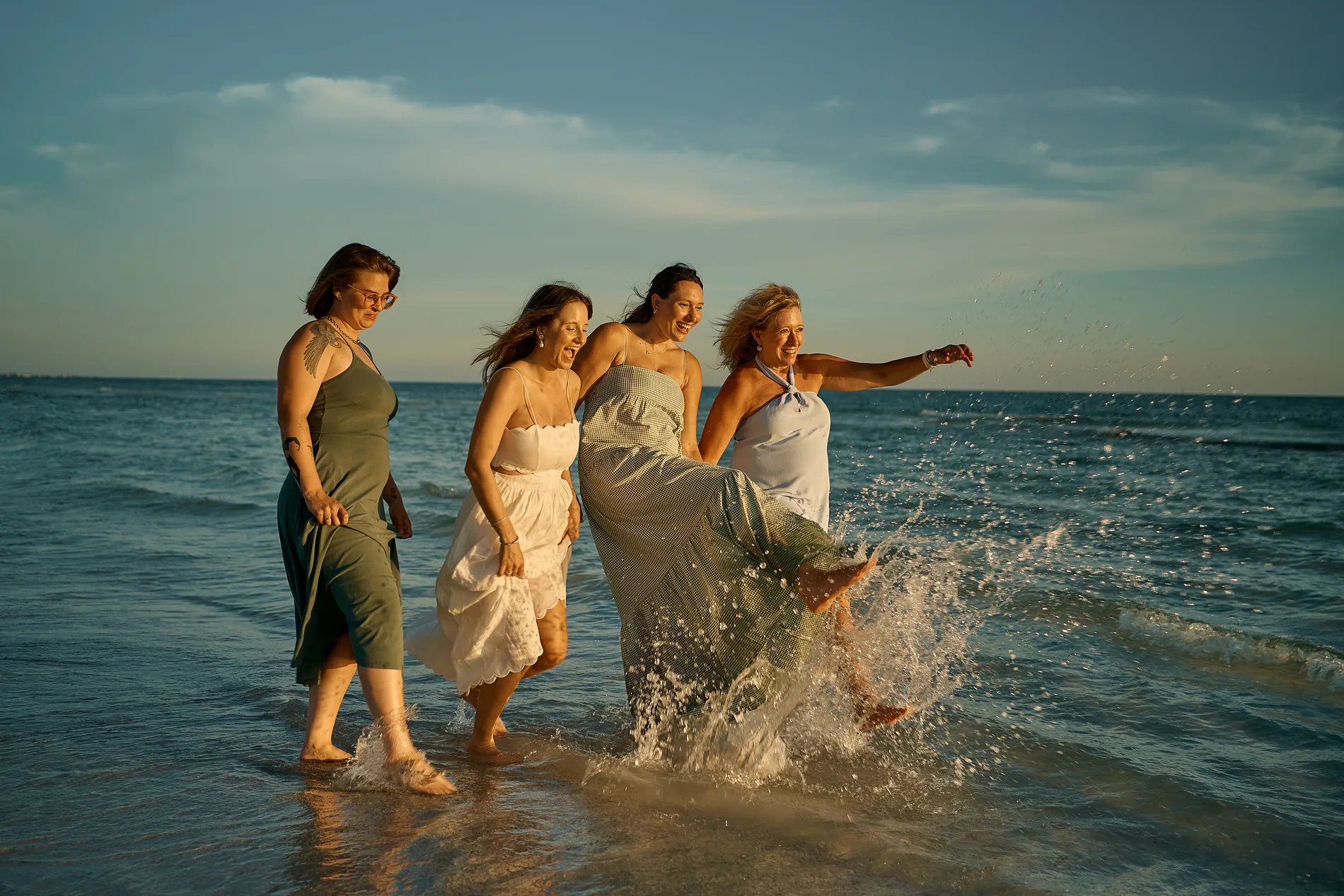 Four women in long flowing dresses in sage, cream, grey-green and pale blue walking through the shallow water, one kicking up a splash, the fabric swinging around their legs